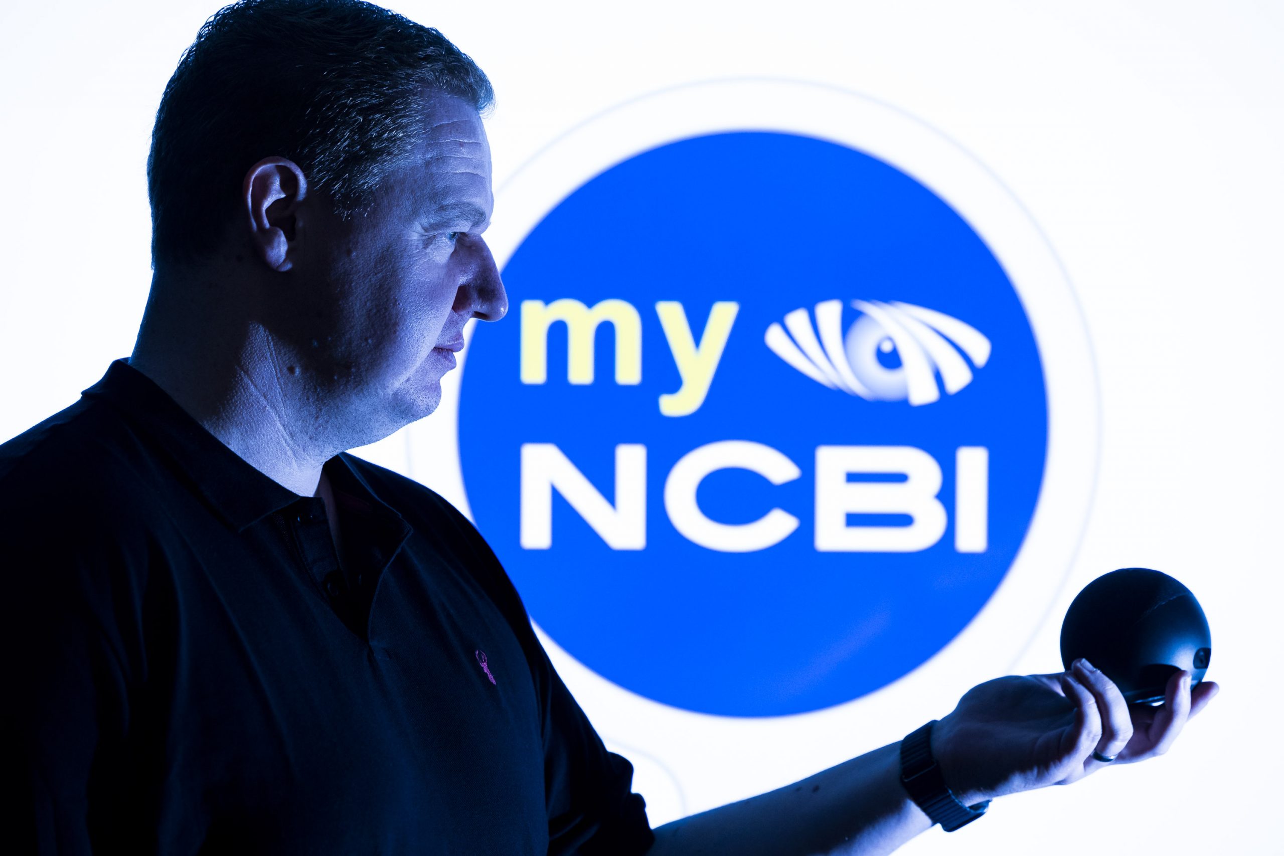 IA Labs CEO, Kyran O'Mahoney, holding an Alexa device in front of the myNCBI logo.