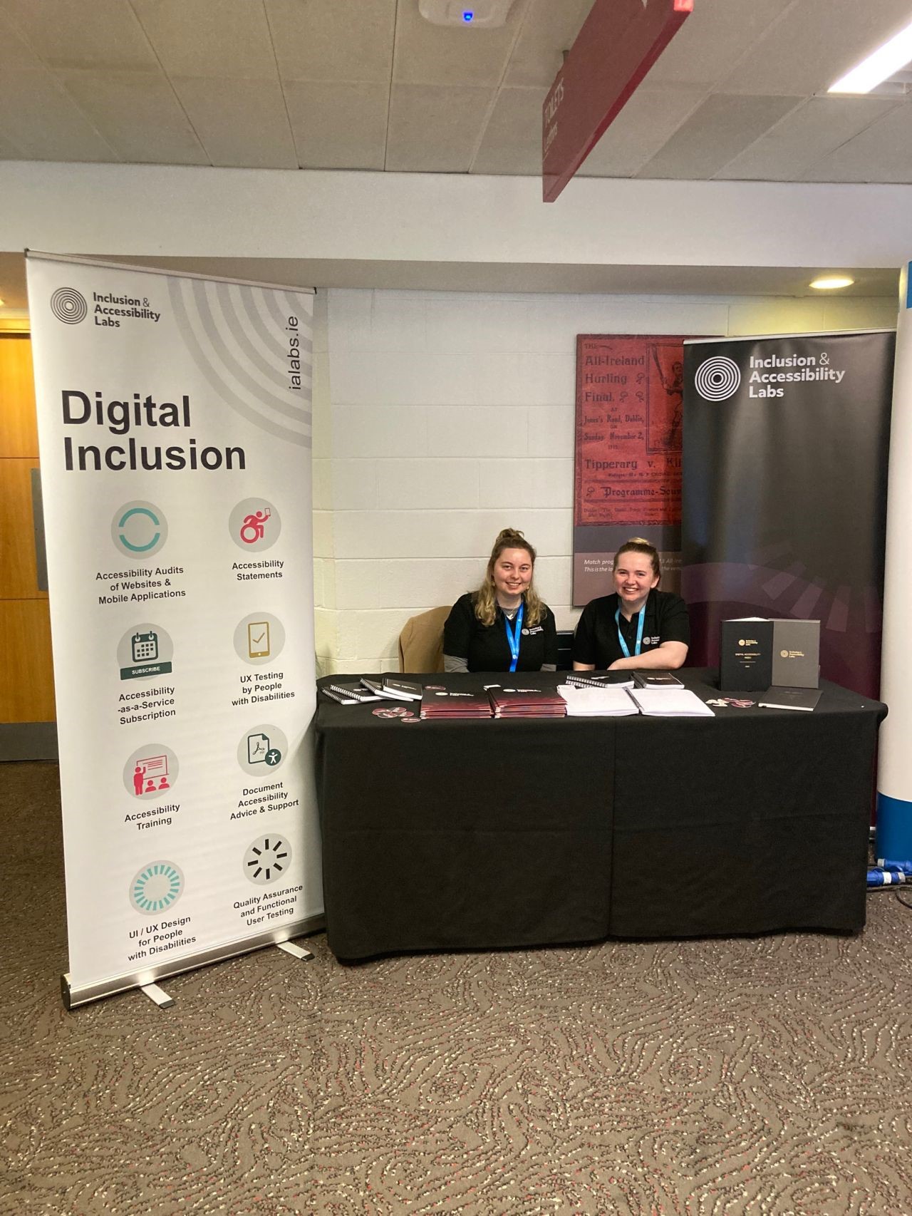 Adela Buliman and Sarah O'Boyle seated at a table surrounded by IA Labs banners