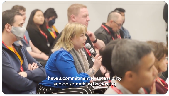 Adela Buliman and Kyran O'Mahoney sitting in the audience of Accessibility Camp Bay Area. Adela is holding a mic to ask a question.