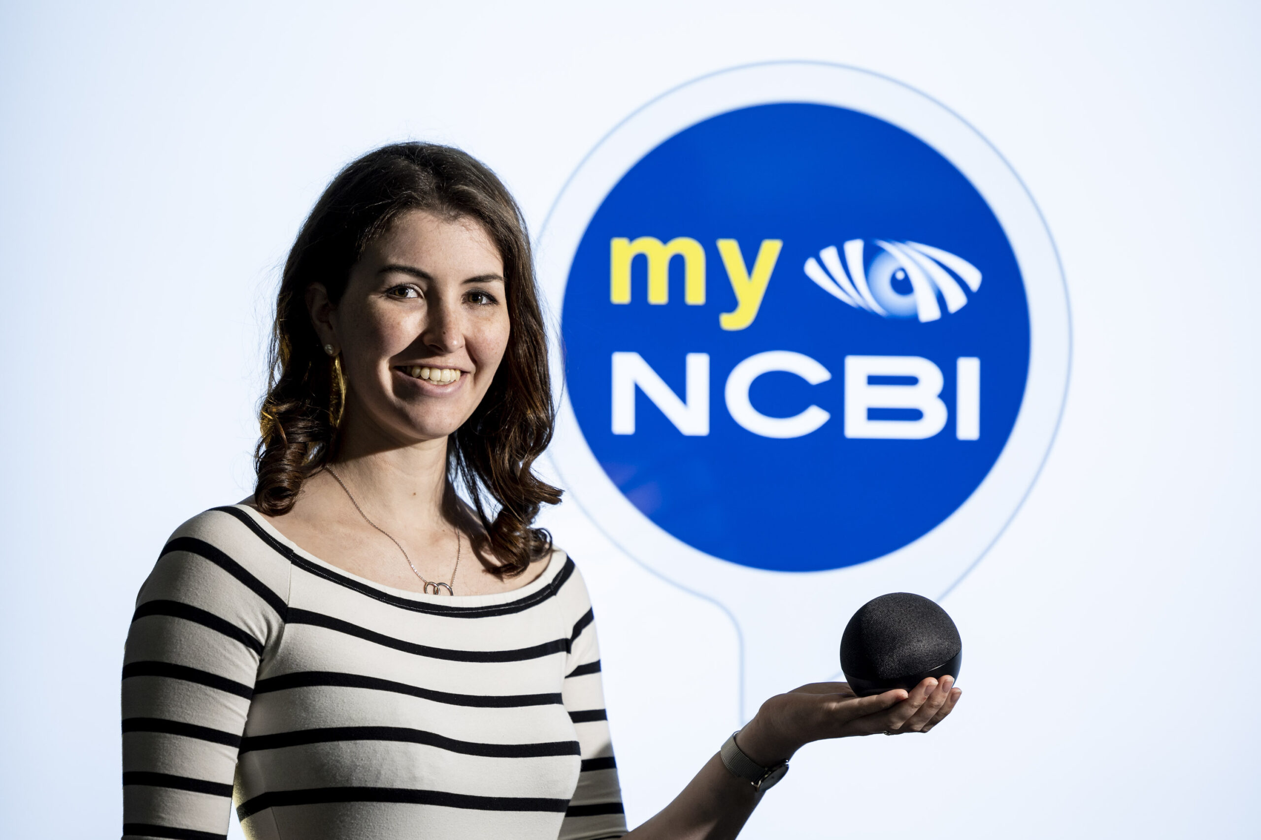 NCBI service user Aine Sullivan holding an Alexa device in front of the myNCBI Smart Hub logo